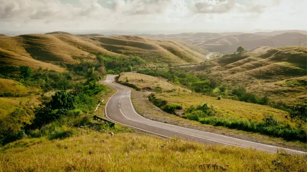 A winding asphalt road cuts through golden, grassy hills under a partly cloudy sky. The landscape is a series of rolling hills and valleys with some green trees and shrubs. The image depicts the natural landscape of Sumba Island, Indonesia.